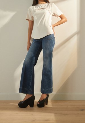 Kobieta ubrana w beżową bluzkę z krótkim rękawem z detalem na przednim panelu, szerokie niebieskie dżinsy z szerokimi nogawkami i czarne platformy z odkrytymi palcami stojąca w pomieszczeniu.