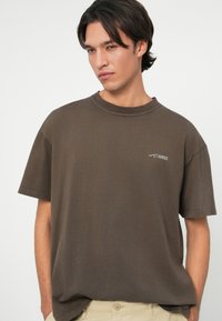 Brązowy t-shirt z krótkim rękawem, z okrągłym dekoltem i małym białym logo na klatce piersiowej. Bawełnowy materiał o gładkiej fakturze i luźnym kroju.
