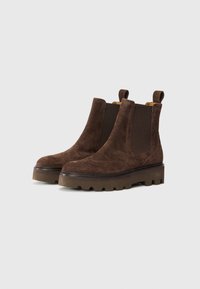 Las botas de tobillo de ante marrón cuentan con paneles laterales elásticos, una suela de goma con cleat y detalles decorativos de brogue en la punta y los lados.