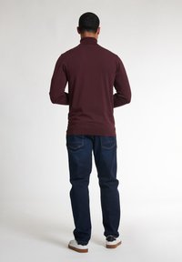 Haut à manches longues bordeaux avec col montant, associé à un jean bleu et des baskets blanches avec des accents noirs, vu de l'arrière.