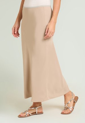 Femme portant une jupe longue beige et des sandales plates argentées à brides, debout devant un fond clair uni.