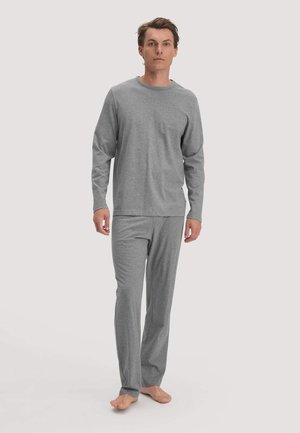 Mann steht barfuß, trägt ein graues Langarmshirt und passende graue Hose vor einfarbigem Hintergrund.
