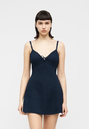 Joven con cabello corto y oscuro, vestida con un vestido sin mangas azul marino con bordes de encaje y un lazo al frente, posando contra un fondo claro y sencillo.