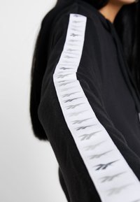 Černá mikina s kapucí a bílým pruhováním na rukávech, zdobená šedými a černými logovými akcenty. Měkká textura, volný střih, s kapucí na šňůrku.