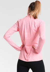 Długosleeve top sportowy w jasnoróżowym kolorze, wykonany z gładkiego, syntetycznego materiału. Posiada wysoki kołnierz oraz sześć tonowych kropek na plecach.