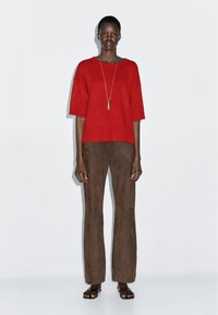 Czerwony dzianinowy sweter z krótkim rękawem, zestawiony z brązowymi zamszowymi spódnicami typu dzwony. Modelka nosi długą złotą naszyjnik z wisiorkiem i czarne sandały.