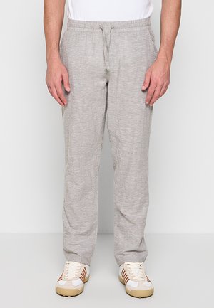 Mann trägt hellgraue Loungebuxe mit Kordelzug und weiße und beige Freizeitsneaker, steht vor einem schlichten hellen Hintergrund.