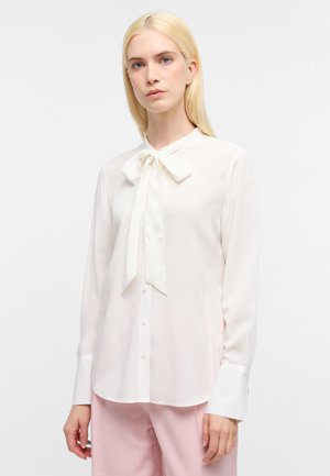 Witte blouse met lange mouwen en een strikbeleg, zachte textuur en knoopsluiting, gecombineerd met lichtroze broek.