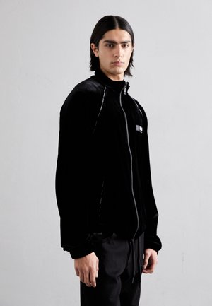 Chaqueta de terciopelo negro con cremallera y alto cuello, que presenta costuras en contraste y bolsillos con cremallera, combinada con pantalones negros.