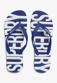 Mėlynos ir baltos šlepetės su tekstūruota padu, turinčios didelį baltą "SUPERDRY" spaudą. Guminiai mėlyni dirželiai su žakardo raštu.
