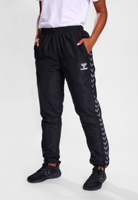 Zwarte joggingsbroek met elastische tailleband, zijzakken en een grijze visgraatpatroon langs de zijkanten. Gecombineerd met zwarte sneakers.