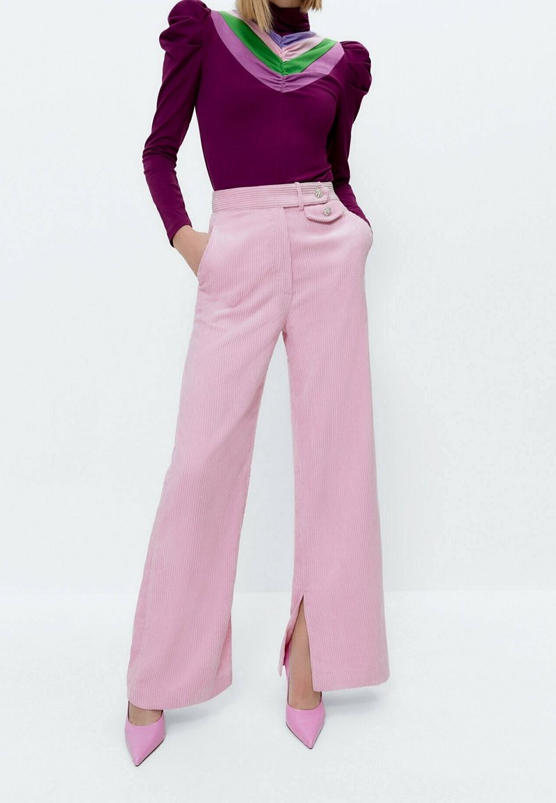 Pantalon large en velours côtelé rose avec fentes latérales, doté de deux boutons sur la ceinture ; associé à un haut ajusté à manches longues violet.