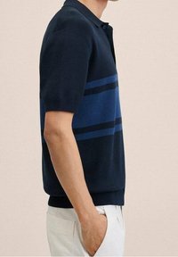 Hombre vistiendo una camiseta polo de manga corta azul marino con rayas horizontales azules y pantalones blancos, de pie con una mano en el bolsillo contra un fondo beige.