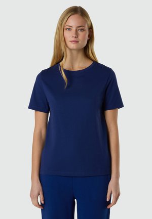 Donna con capelli lunghi e biondi che indossa una maglietta a maniche corte blu navy semplice con scollo rotondo e pantaloni abbinati, in piedi contro uno sfondo grigio chiaro.