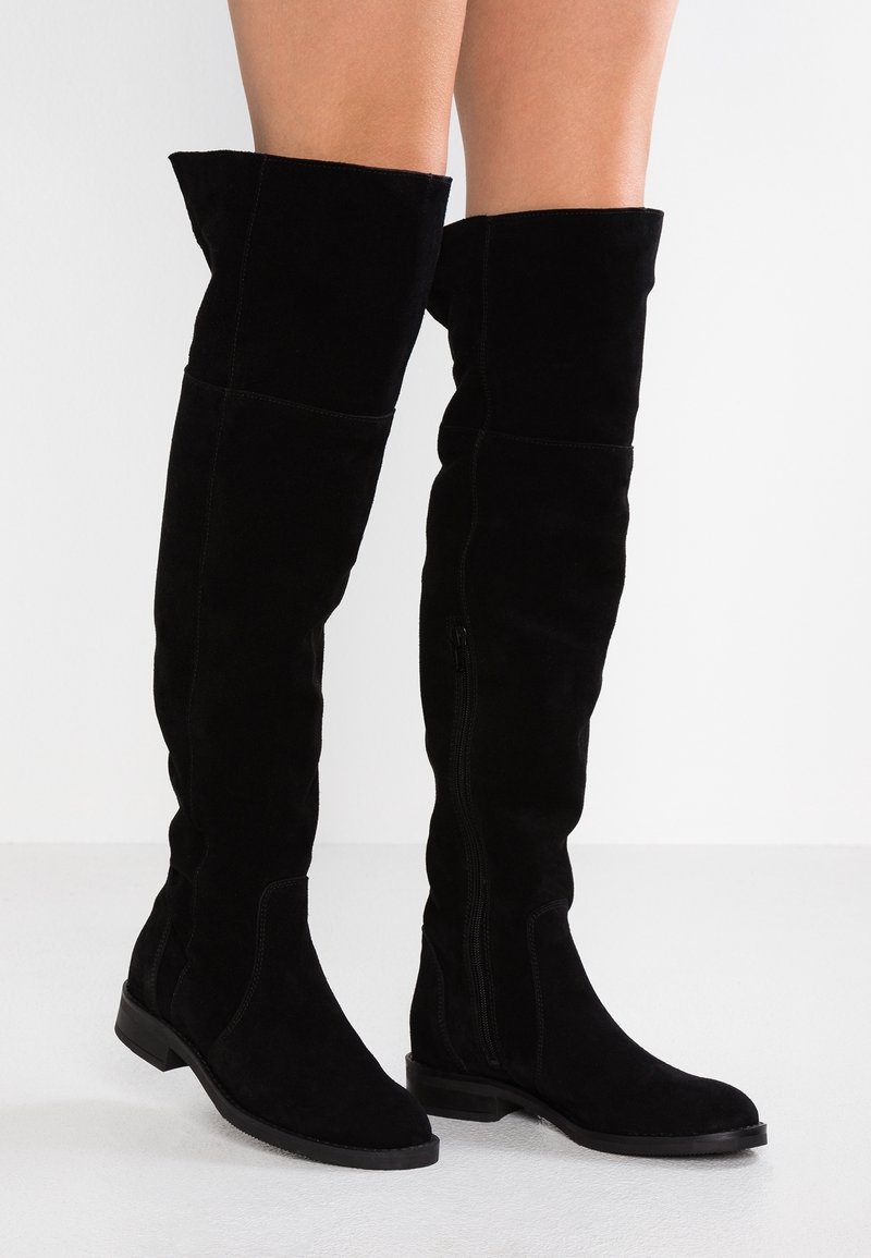 Bottes en daim noir montant au-dessus du genou avec une semelle plate. Elles présentent une silhouette élancée, une fermeture éclair latérale et un revers plié en haut.