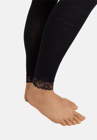 Zwarte geribde panty met een doorzichtige kantafwerking bij de enkel, versierd met bloemmotieven en golvende randen. Voet is zichtbaar.