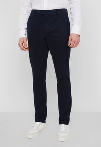 Marineblaue Baumwollhose mit schmalem Schnitt, mit flachem Bund, einem Knopfverschluss und seitlichen Taschen. Getragen mit weißen Sneakern.
