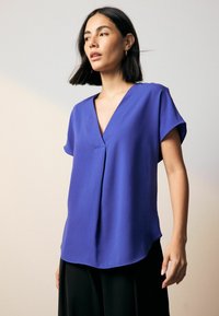 Hladká blouse s krátkým rukávem a výstřihem do V v tmavě modré barvě. Obsahuje jemný záhyb vpředu a zakřivený lem.