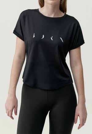 Frau trägt schwarzes, kurz geschnittenes T-Shirt mit hochgekrempelten Ärmeln und minimalistischen weißen Buchstaben auf der Brust, kombiniert mit schwarzen Leggings.
