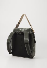 Sac étanche vert avec un dos en mesh noir texturé, doté d'une sangle réglable beige, de poignées robustes et d'un design minimaliste.