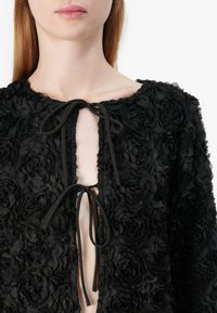 Vêtement noir orné de roses en tissu, avec un lien de fermeture au cou et un design à l'avant ouvert. Apparence texturée et superposée.