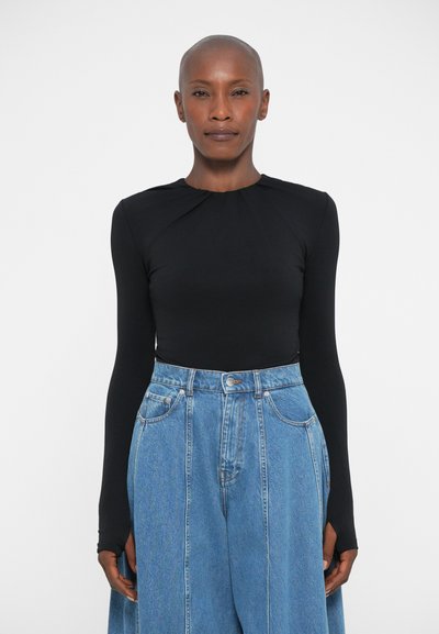 Mujer con la cabeza rapada, vestida con una camiseta ajustada de manga larga negra y unos pantalones vaqueros de pierna ancha de talle alto en un fondo blanco liso.