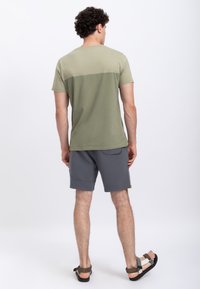 T-shirt verde oliva con un blocco di colore orizzontale, abbinato a pantaloni corti grigi scuri con tasca posteriore e cuciture piatte. Sono indossate sandali piatti.