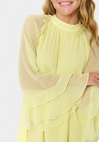 Flowy yellow chiffon dress with a high neckline and long, layered bell sleeves. Features soft texture and a lightweight design.
