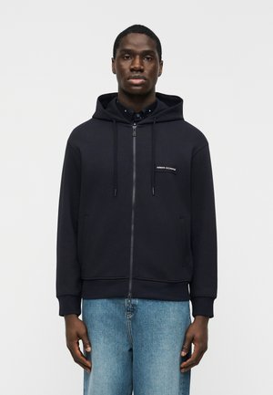 Moški, ki nosi črno jopa z zadrgo z napisom "Armani Exchange" na prsih in modre kavbojke, stoji pred preprostim belim ozadjem.