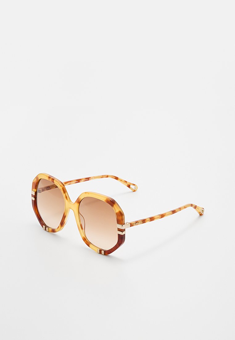 Übergroße achteckige Schildkrötensonnenbrille mit braunen Verlaufslinsen und dünnen, passenden Bügeln, auf einer weißen Oberfläche platziert.