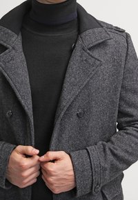 Cappotto grigio in lana doppiopetto con tessuto texturizzato, ampi rever e dettagli con bottoni, indossato sopra un maglione a collo alto nero.