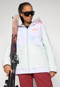Giacca da sci in colori pastello chiari con motivo testurizzato, chiusura con zip e logo. Abbinata a pantaloni da sci scuri e attrezzatura da sci.