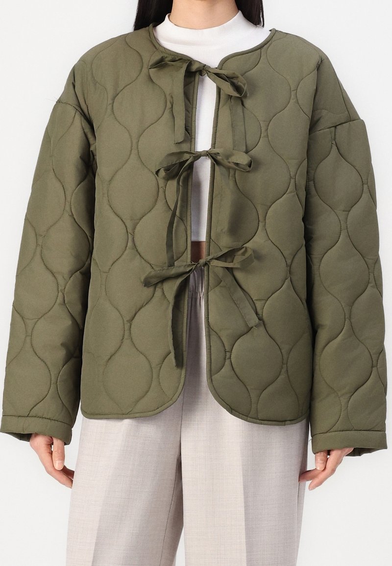 Veste matelassée vert olive avec un col arrondi, trois fermetures par nœuds et un motif texturé, portée avec un pantalon clair.