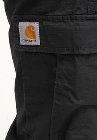Svart tyg med en texturerad mönster har en vit etikett med en orange logotyp. Designen inkluderar en synlig ficka och sömnadsdetaljer.