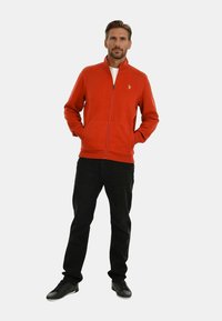 Rote Fleece-Zip-Jacke mit hohem Kragen, kombiniert mit schwarzen Hosen und schwarzen Sneakern. Besitzt ein kleines Logo auf der Brust.