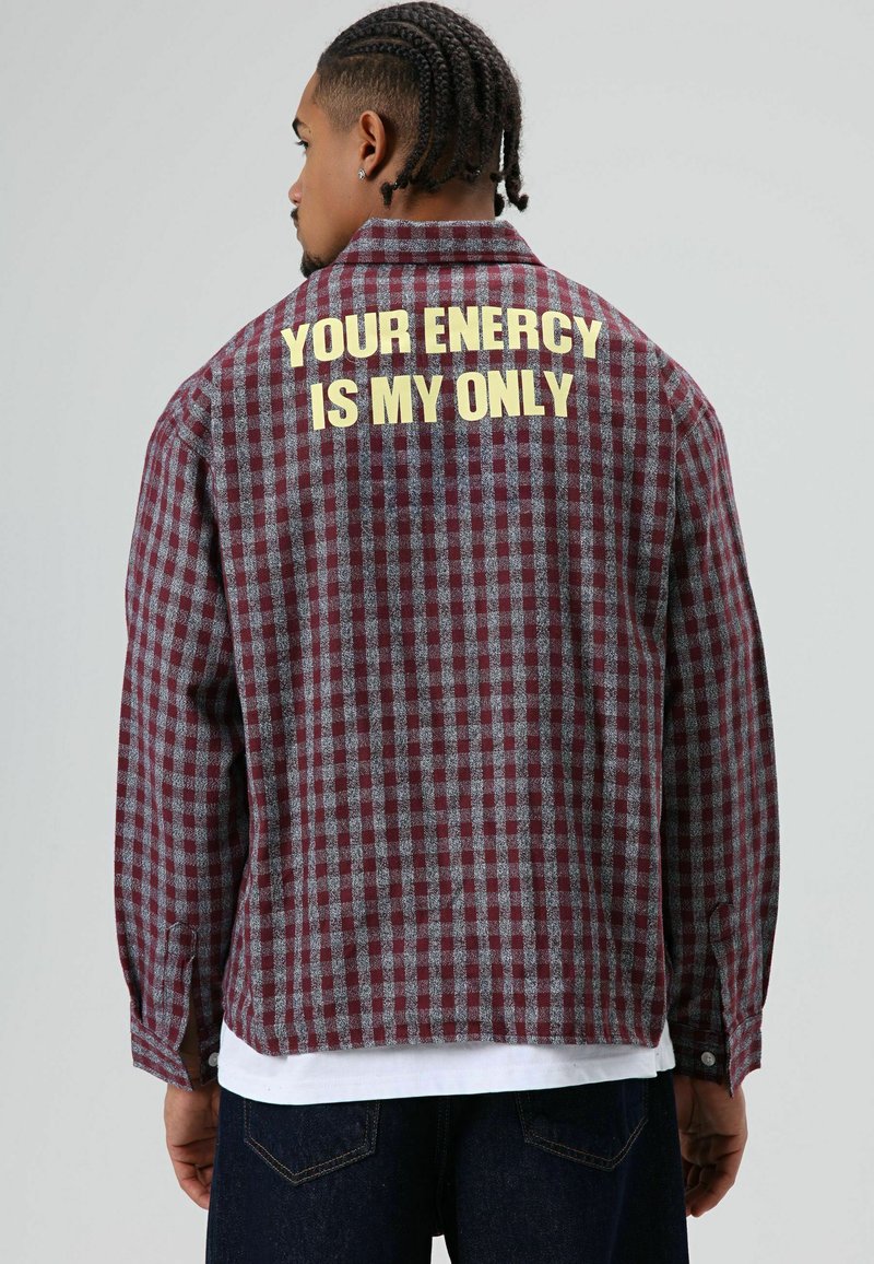Rot-grau kariertes Flanellhemd mit langen Ärmeln, auf dem Rücken groß gelber Text mit der Aufschrift "DEINE ENERGIE IST MEIN EINZIGES".