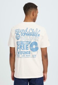 Wit katoenen T-shirt met blauwe tekst op de achterkant ter promotie van een evenement, met grafische elementen waaronder een vinylplaat en een handillustratie.