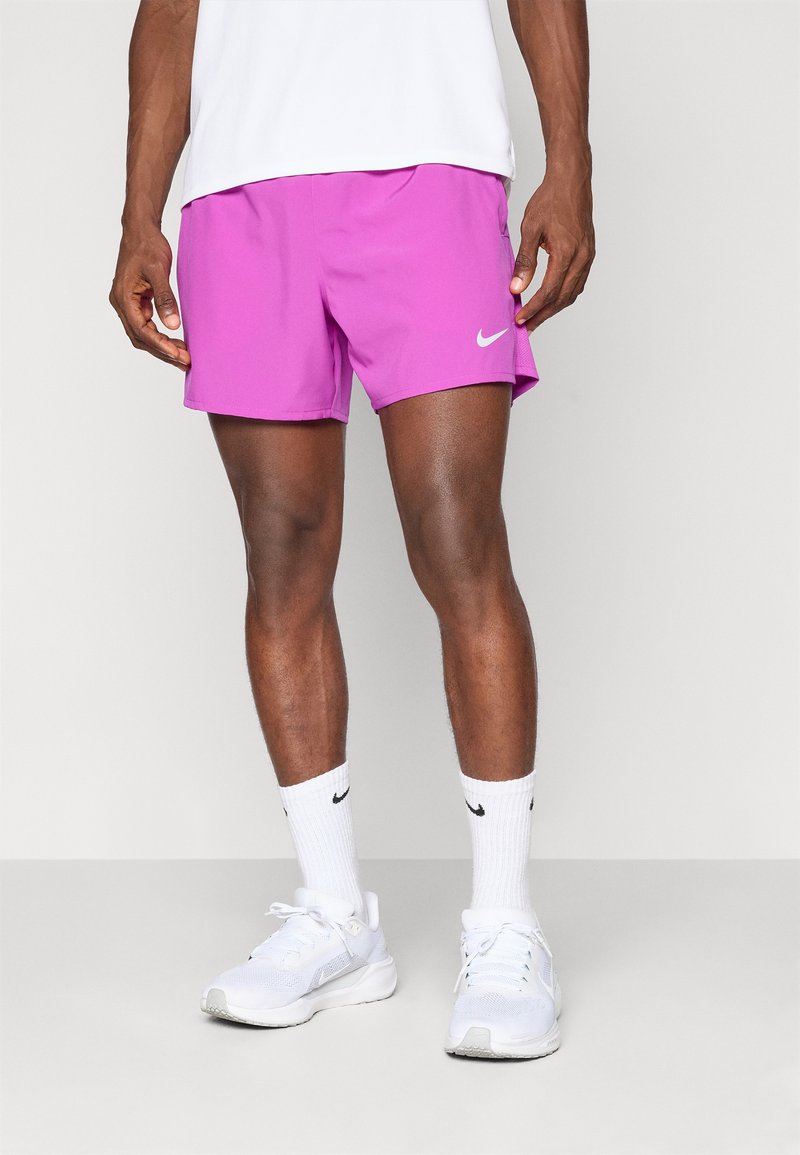 Homme portant un short de sport rose vif, des chaussettes blanches Nike et des chaussures de course blanches, debout devant un fond gris clair.