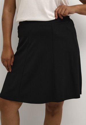 Personne avec un teint de peau moyen portant une jupe noire longueur genou et un haut blanc, ajustant la taille d'une main.