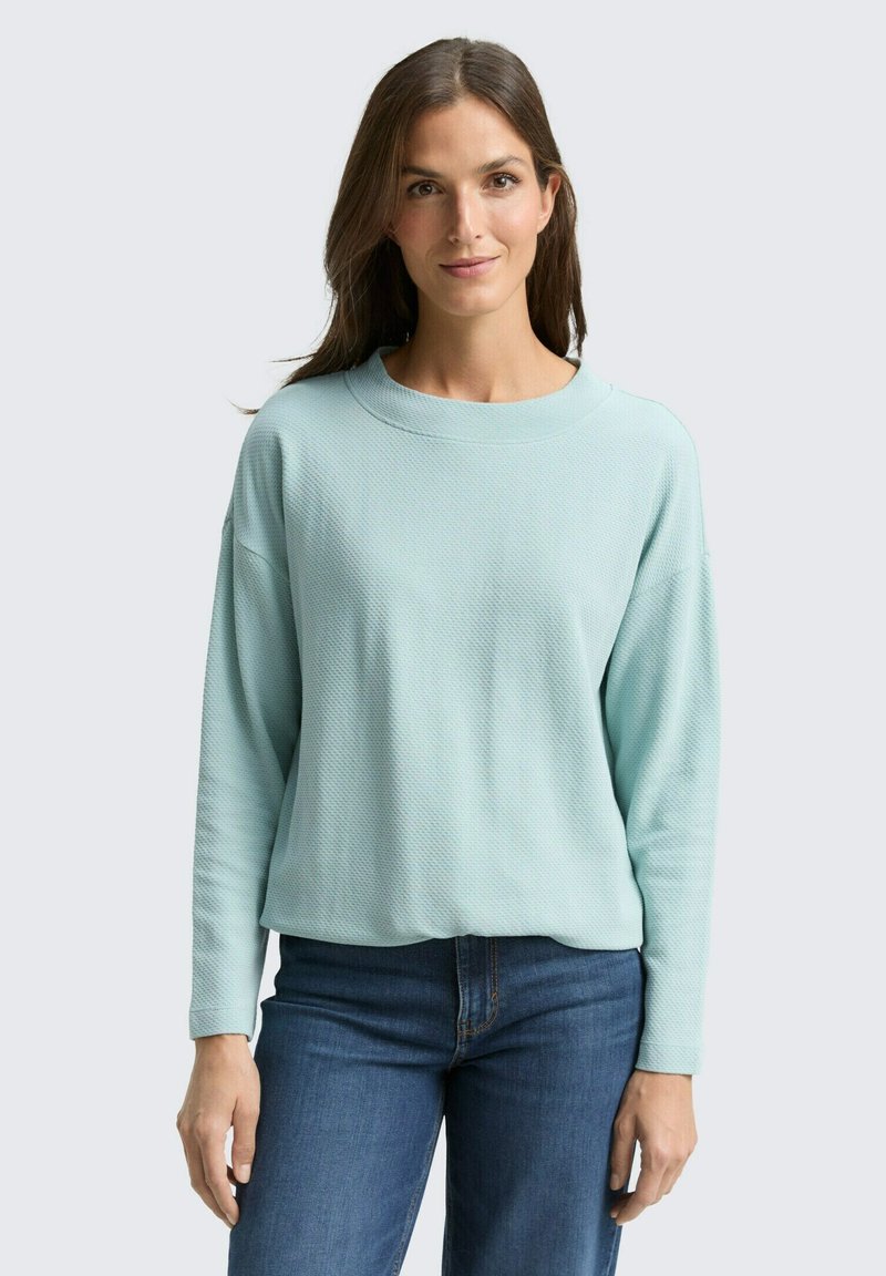 Himmelblaues, strukturiertes Sweatshirt mit weitem Halsausschnitt und überschnittenen Schultern, kombiniert mit blauen Jeans. Einfaches, lässiges Design.