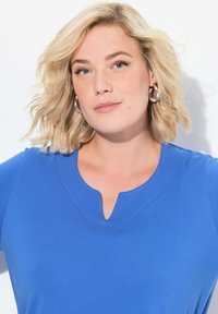 Blondýna s vlnitými vlasy, oblečená v modrém topu s výstřihem a stříbrnými kruhovými náušnicemi, na bílém pozadí.
