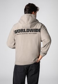 Beige Fleece-Kapuzensweatshirt mit einem großen schwarzen "WORLDWIDE"-Schriftzug auf dem Rücken. Mit verstellbarer Kapuze und lockerem Schnitt. Kombiniert mit schwarzen Hosen.