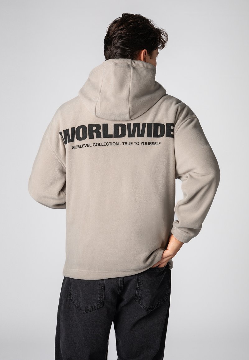 Beige Fleece-Kapuzensweatshirt mit einem großen schwarzen "WORLDWIDE"-Schriftzug auf dem Rücken. Mit verstellbarer Kapuze und lockerem Schnitt. Kombiniert mit schwarzen Hosen.
