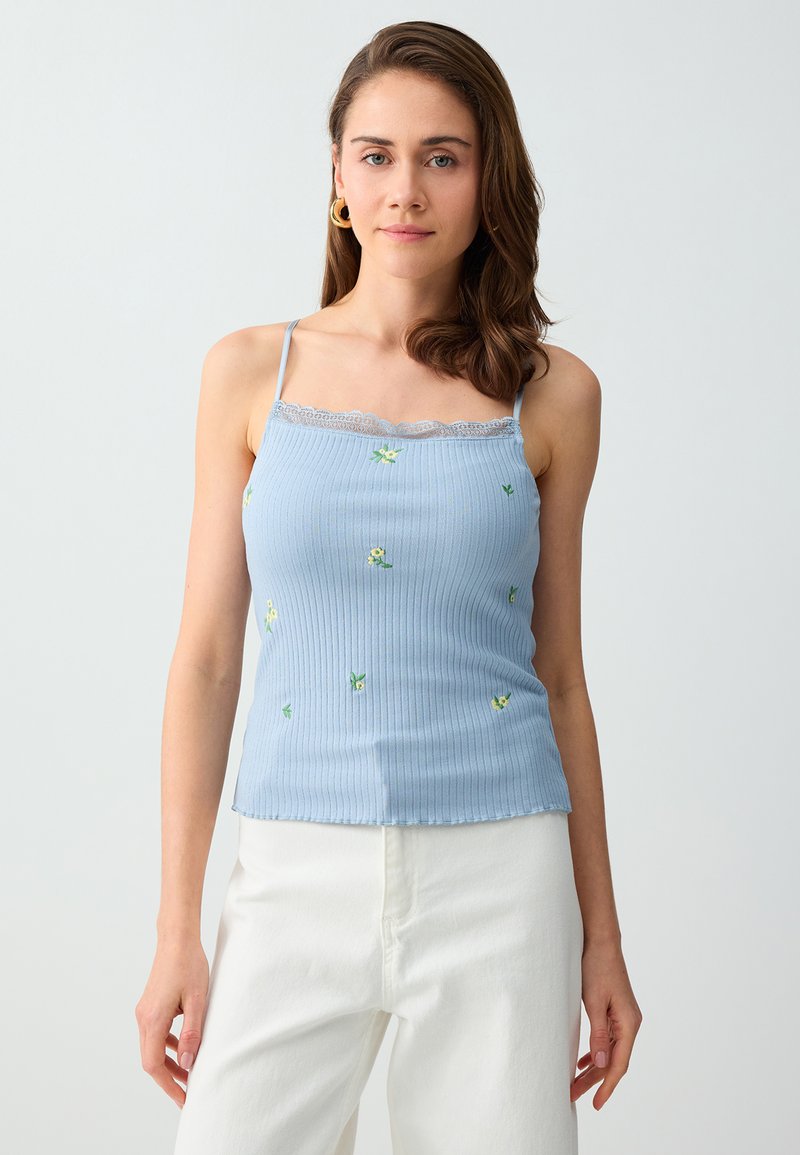 Femme aux cheveux bruns mi-longs portant des boucles d’oreilles créoles dorées, un caraco fleuri bleu clair avec bordure en dentelle et un pantalon blanc sur un fond uni.