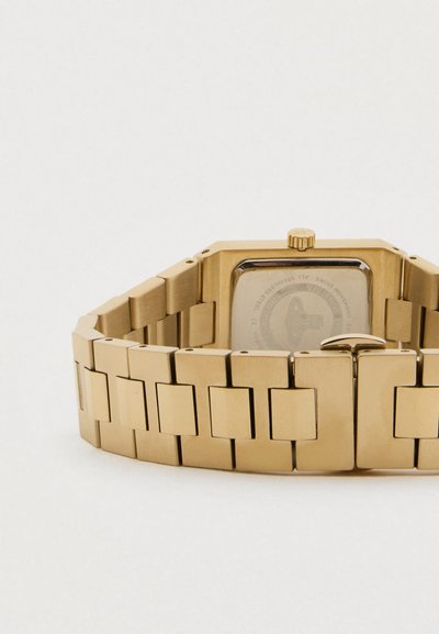 Bracelete de relógio em metal dourado com fecho, mostrando a parte de trás de uma caixa de relógio quadrada sobre um fundo branco.