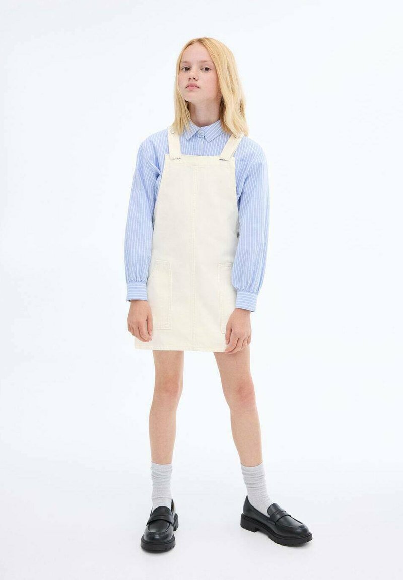 Witte denim tuinbroekjurk met voorzakken, gedragen over een lichtblauw gestreept shirt. Gecombineerd met grijze sokken en zwarte instap schoenen.