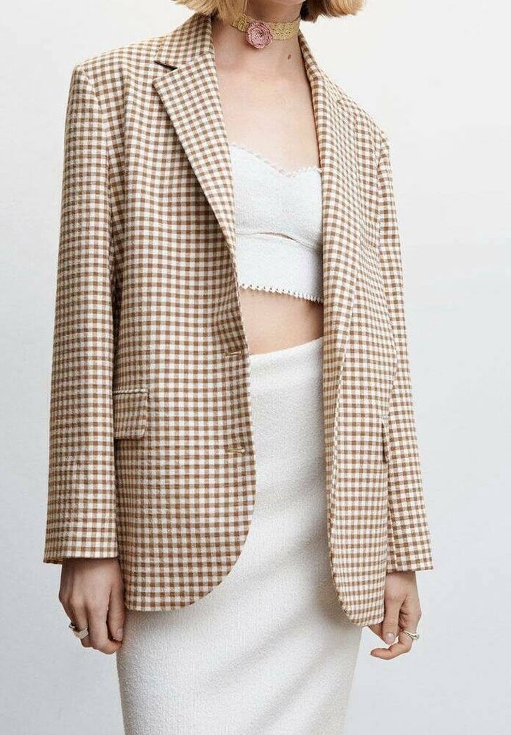 Femme portant un blazer à carreaux marron et blanc, un crop top blanc texturé, une jupe blanche ajustée et un collier ras du cou beige avec un détail de fleur rose.