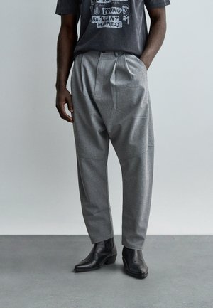 Hombre con pantalones grises sueltos plisados, una camiseta gráfica oscura y botines de cuero negro, de pie sobre un suelo gris.