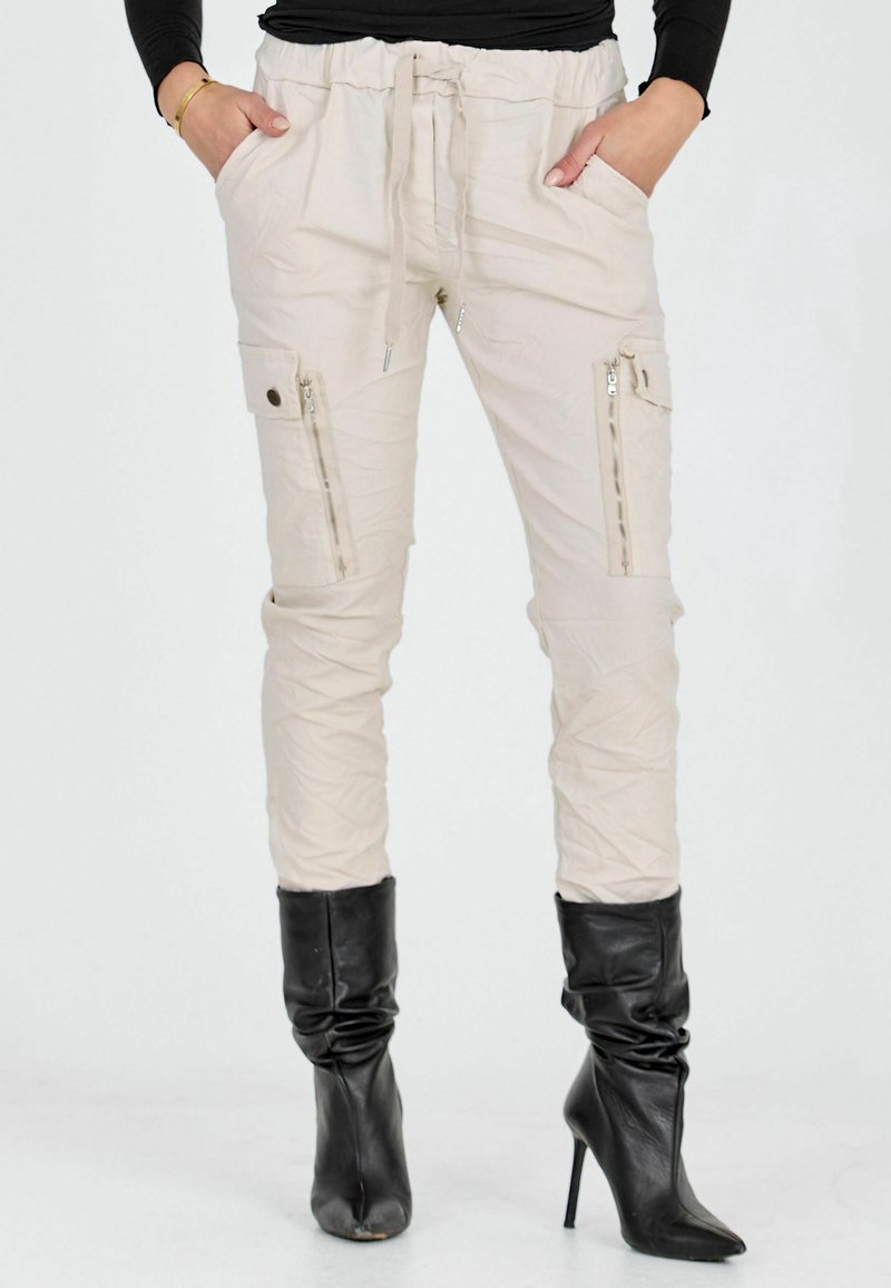 Személy bézs színű cargo nadrágot visel cipzáras zsebekkel és fekete, magas sarkú, laza csizmával, kezei a zsebekben, fehér háttér előtt áll.