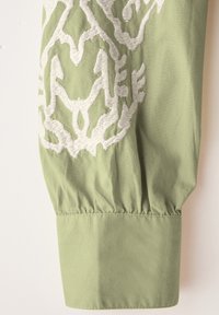 Blusa di cotone verde con maniche, decorata con motivo floreale ricamato bianco. Texture liscia, polsino arricciato alla fine, design sartoriale.